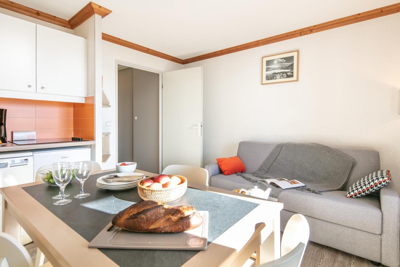 Une cuisine moderne avec des meubles blancs, une table à manger sur laquelle est posée une miche de pain, et un canapé gris agrémenté d’un coussin rouge.