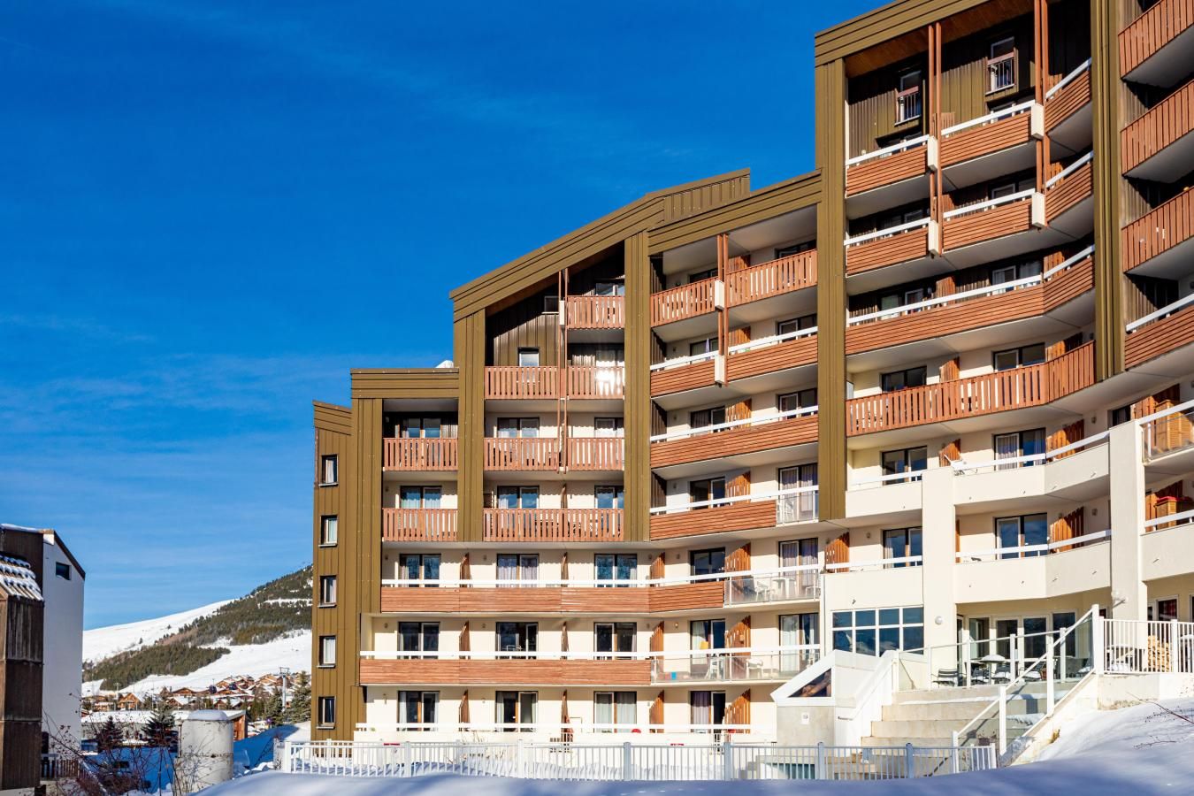 Un hôtel à plusieurs étages doté de balcons, avec une piste de ski en arrière-plan et un ciel bleu dégagé.