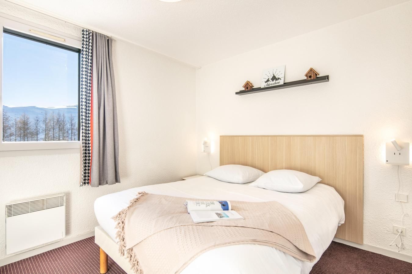 A hotel room with a bed, a window with curtains, a wall-mounted shelf with decorative items, and a wall socket.