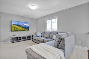 Ein modernes Wohnzimmer mit einer Eckcouch, einem Flachbildfernseher an der Wand, einem Fenster mit Blick und einer Deckenleuchte.