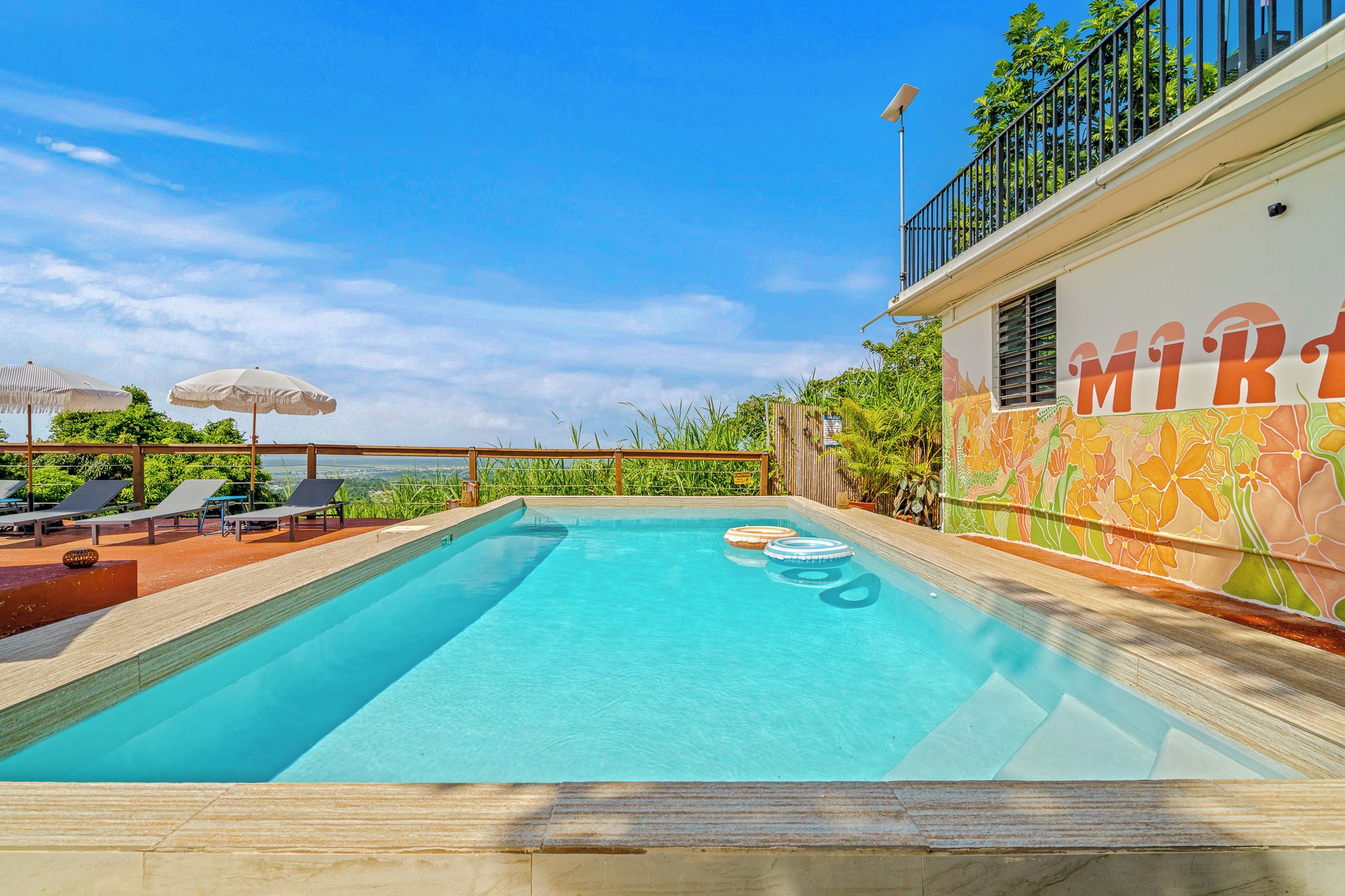 Piscina com espreguiçadeiras e guarda-sóis, rodeada por um deck de madeira e um mural colorido.