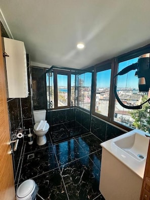 Ein modernes Badezimmer mit Toilette, Waschbecken und Duschbereich, ausgestattet mit großen Fenstern und Blick auf die Stadtlandschaft.