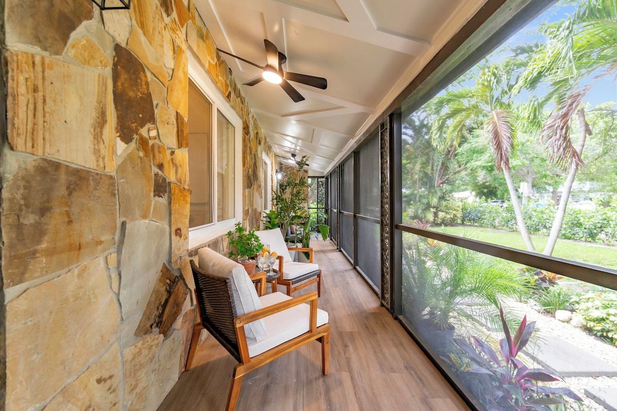 A covered outdoor seating area with stone walls, wooden furniture, and a view of tropical vegetation.
