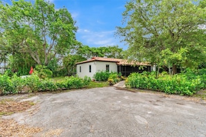 A single-story house with a tiled roof, surrounded by lush greenery and a paved driveway. - Tropical Oasis - 3BR, 10 Min Bal Harbour- Parking (Biscayne Park)