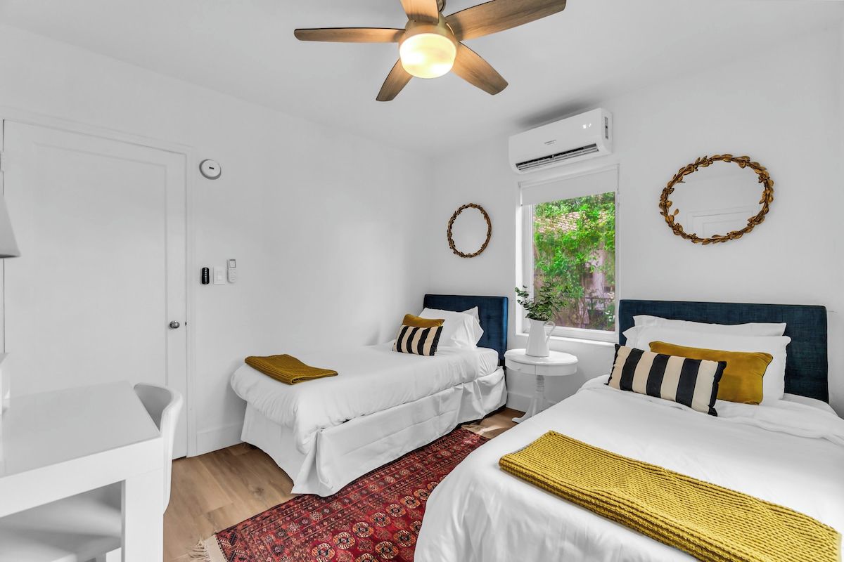 A hotel room with two beds, a ceiling fan, an air conditioning unit, a window with a view of greenery, and a red patterned rug.
