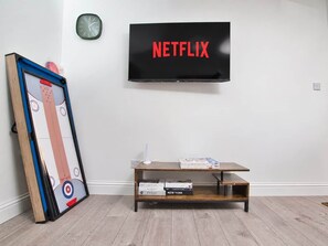 A room with a television mounted on the wall displaying 'NETFLIX,' a wooden coffee table with books, and a framed picture leaning against the wall.
