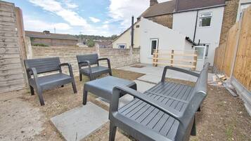 Un espace extérieur aménagé avec des bancs et une table, entouré d’une clôture en bois et d’un bâtiment à la façade blanche.