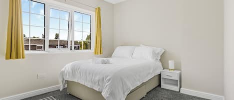 A bedroom with a bed, a nightstand, a lamp, a window with curtains, and a view of buildings outside.