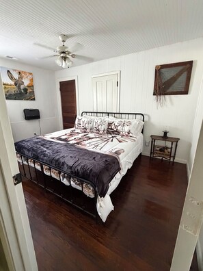 Un dormitorio con cama, ventilador de techo, puerta de madera, una mesita y un cuadro colgado en la pared.