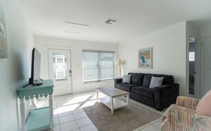 Een woonkamer met een bank, een salontafel, een televisie op een meubel en een raam met jaloezieën.