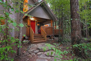 A cabin with a wooden staircase leading to a red door, surrounded by trees and a stone pathway. - Cedar Hollow Mt Baker Cabin with Hot Tub and Stone Fireplace (Deming)