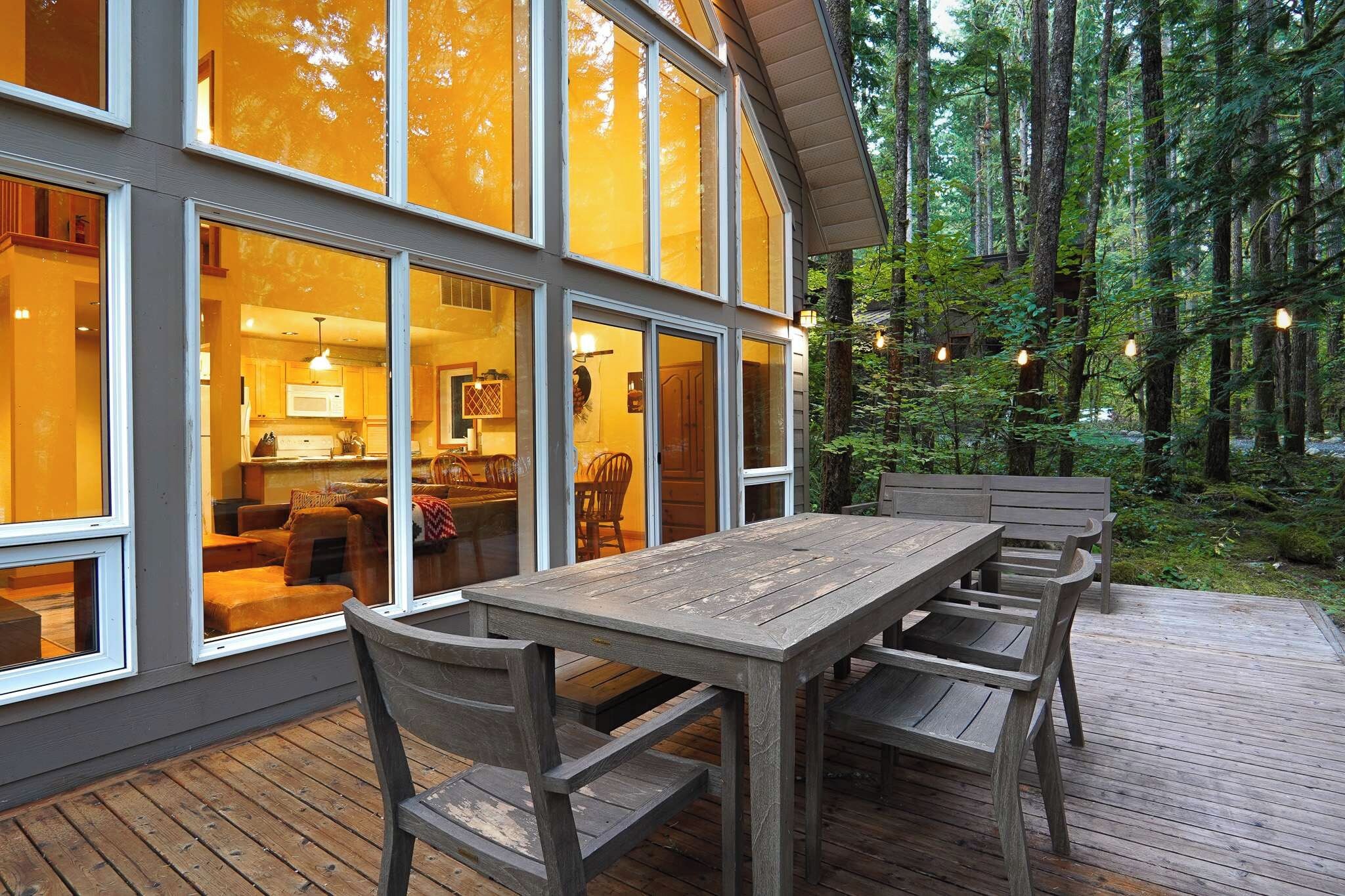 A wooden deck with a dining table and chairs, surrounded by a forested area.