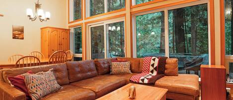 A spacious living room with a large brown leather sectional sofa, a wooden coffee table, and a dining area with wooden chairs and a cabinet.
