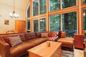 A spacious living room with a large brown leather sectional sofa, a wooden coffee table, and a dining area with wooden chairs and a cabinet. - Cedar Hollow Mt Baker Cabin with Hot Tub and Stone Fireplace (Deming)