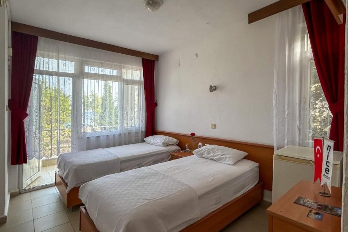 A hotel room with two single beds, a wooden headboard, a small bedside table with a lamp, a nightstand, and a window with curtains.