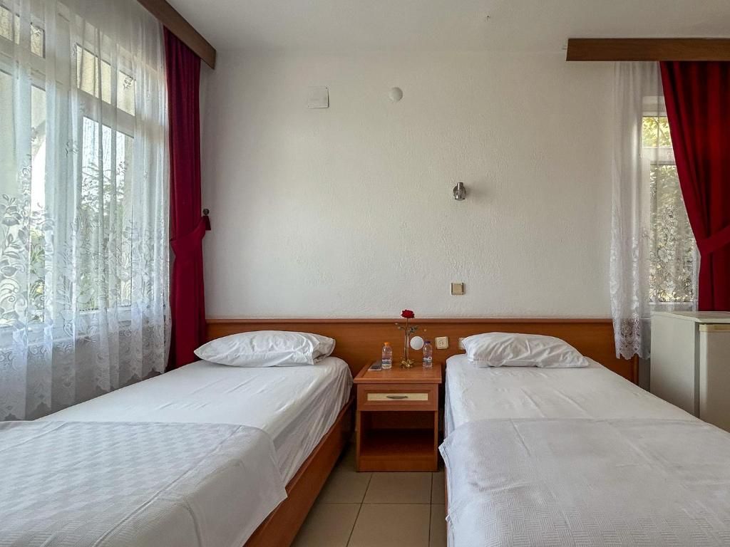 Habitación de hotel con dos camas individuales, una mesita de noche de madera con un espejo y una rosa roja, y una ventana con cortinas de gasa.