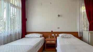 Habitación de hotel con dos camas individuales, una mesita de noche de madera con un espejo y una rosa roja, y una ventana con cortinas de gasa.