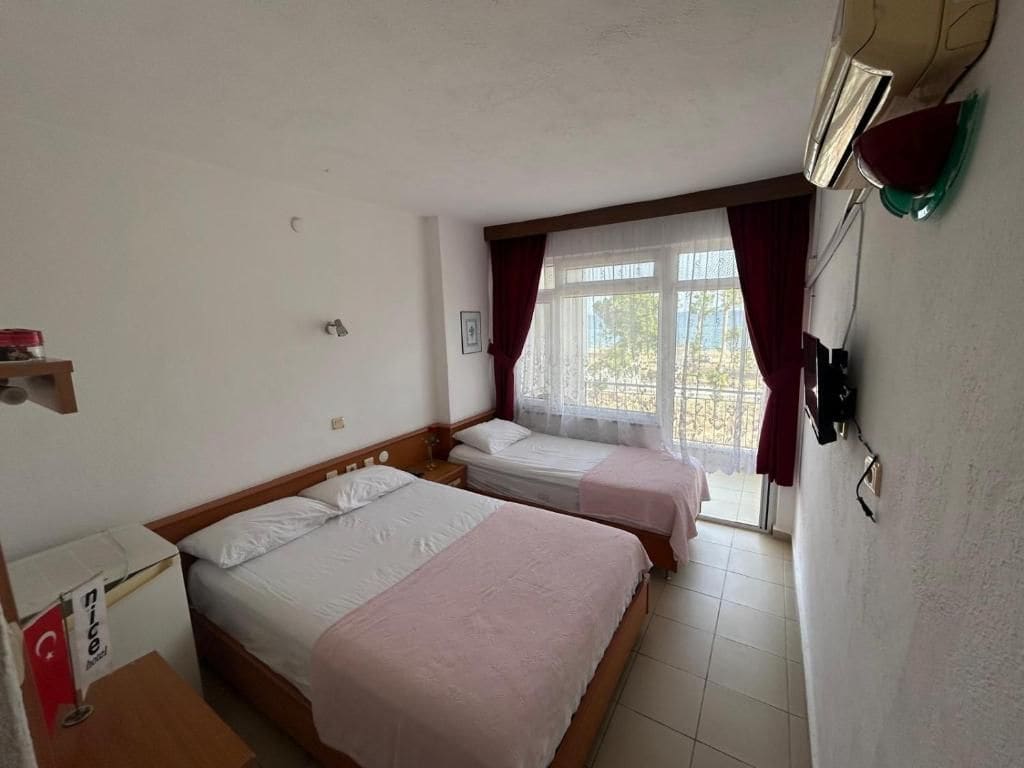 Habitación de hotel con dos camas, una ventana con cortinas, un aire acondicionado instalado en la pared y una mesita con lámpara.