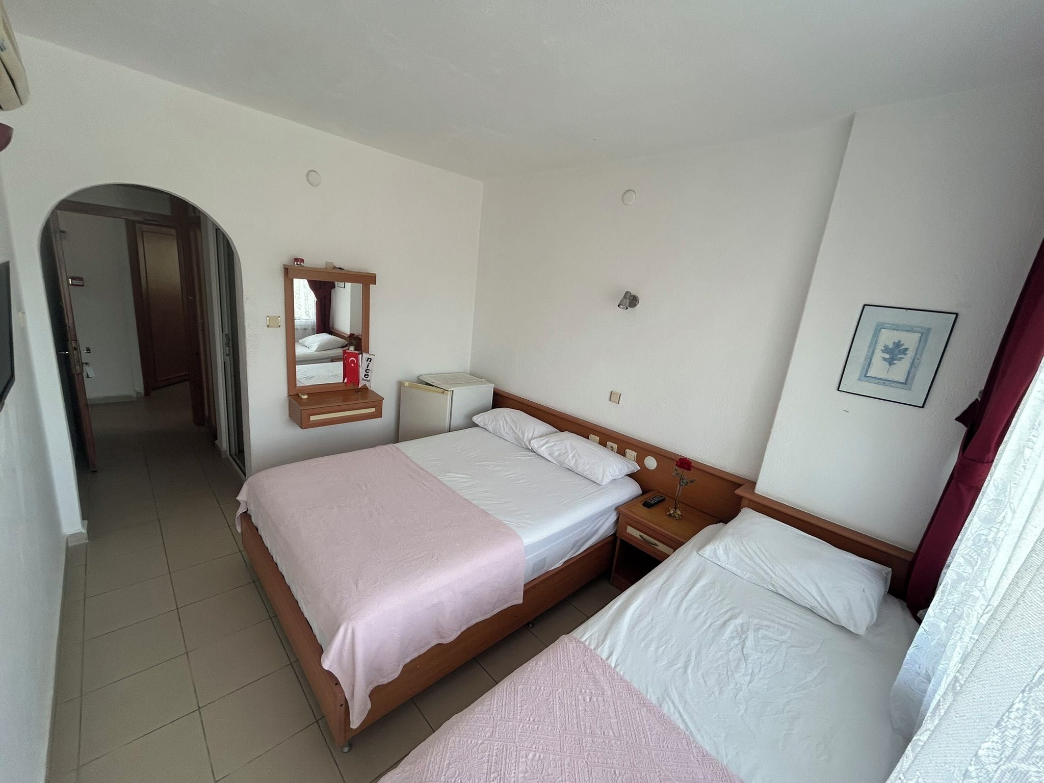 A hotel room with a double bed, a bedside table with a lamp, a mirror, a small bedside table with a remote control, and a picture on the wall.