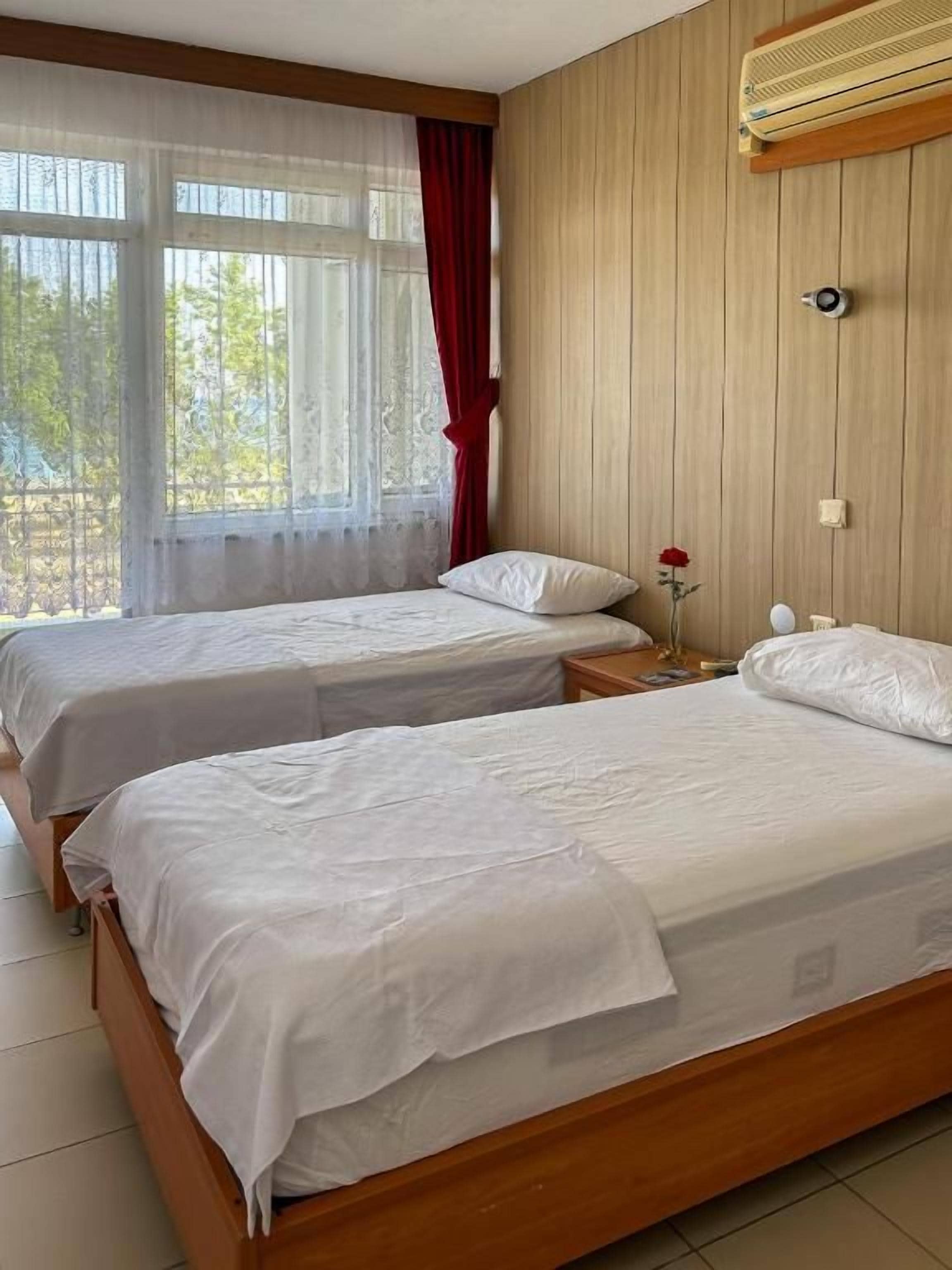 Una habitación con dos camas de madera, una ventana con cortinas de encaje, un panel de pared de madera y una mesita de noche con una lámpara.