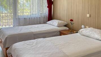 Una habitación con dos camas de madera, una ventana con cortinas de encaje, un panel de pared de madera y una mesita de noche con una lámpara.