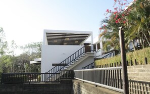 A modern building with a staircase leading to a white upper level, surrounded by greenery and a clear sky.