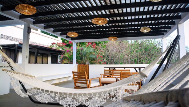 Un hamac orné d’une bordure blanche en crochet, des chaises en bois et un plafond avec des poutres noires et des luminaires suspendus.
