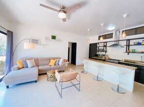 A modern living room with a sofa, a dining area with bar stools, a kitchen with a stove, and a ceiling fan.