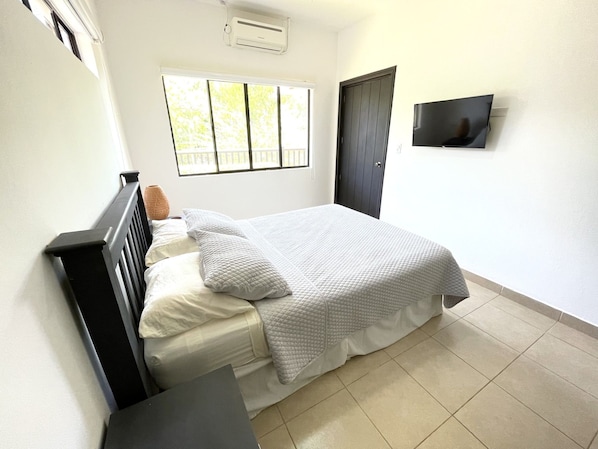 A bedroom with a bed, a bedside table, a window, an air conditioning unit, and a door.