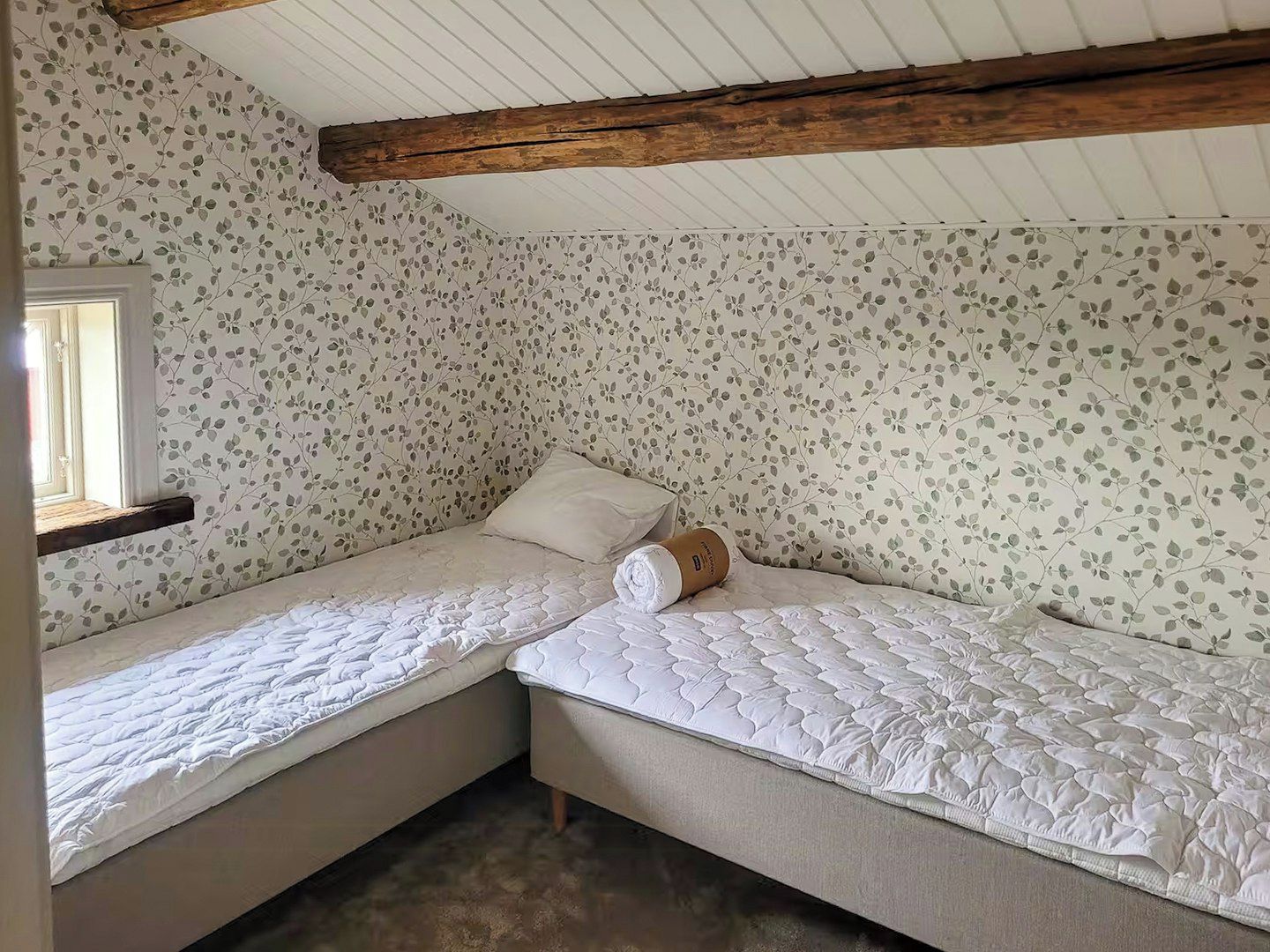 Une chambre avec deux lits simples, une poutre en bois au plafond et un papier peint à motifs floraux.