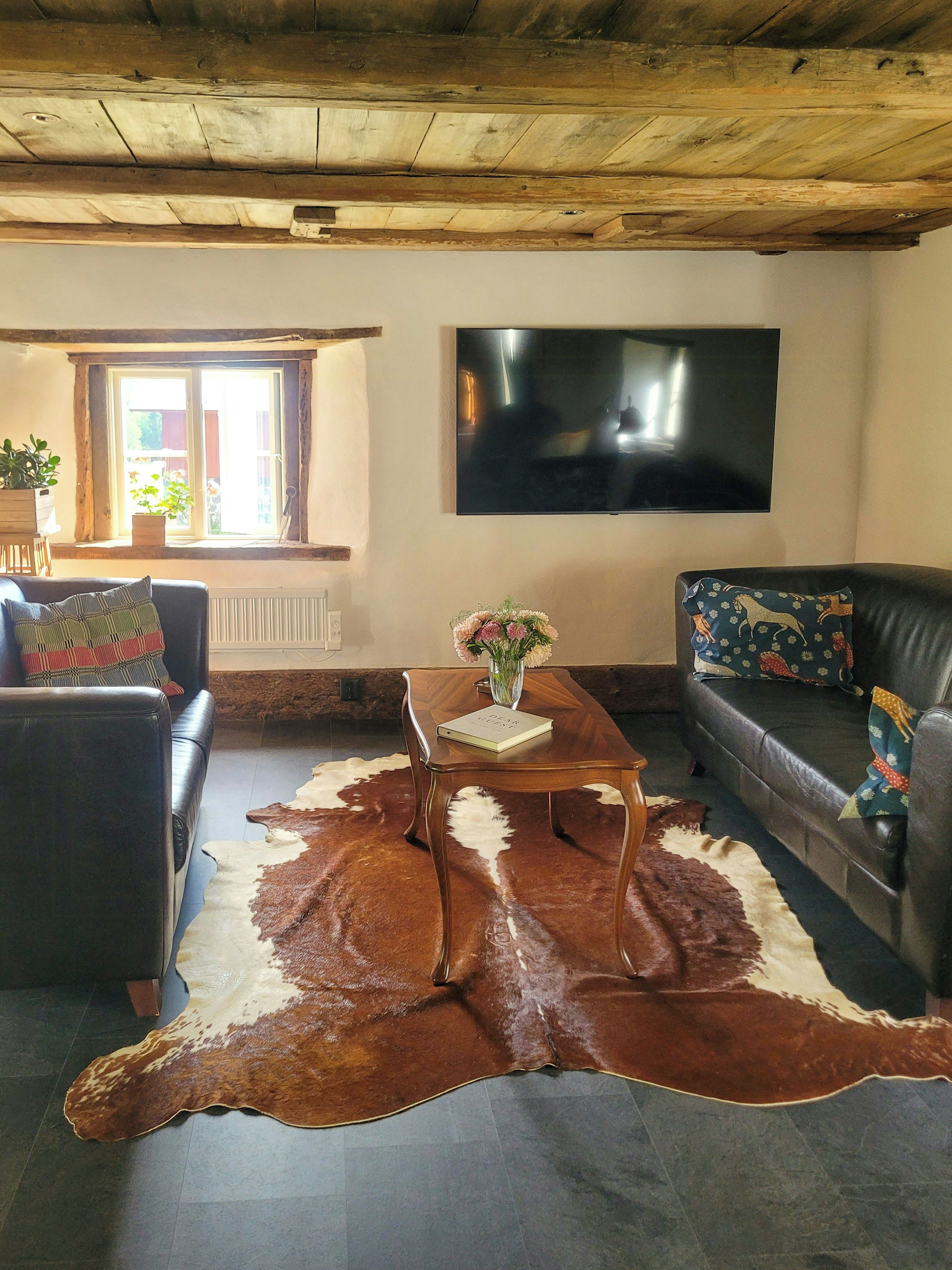 Een woonkamer met een houten plafond, een flatscreen televisie aan de muur, een leren bank, een bijzettafel met een boek en vaas, en een koehuidkleed.