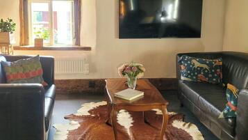 Een woonkamer met een houten plafond, een flatscreen televisie aan de muur, een leren bank, een bijzettafel met een boek en vaas, en een koehuidkleed.