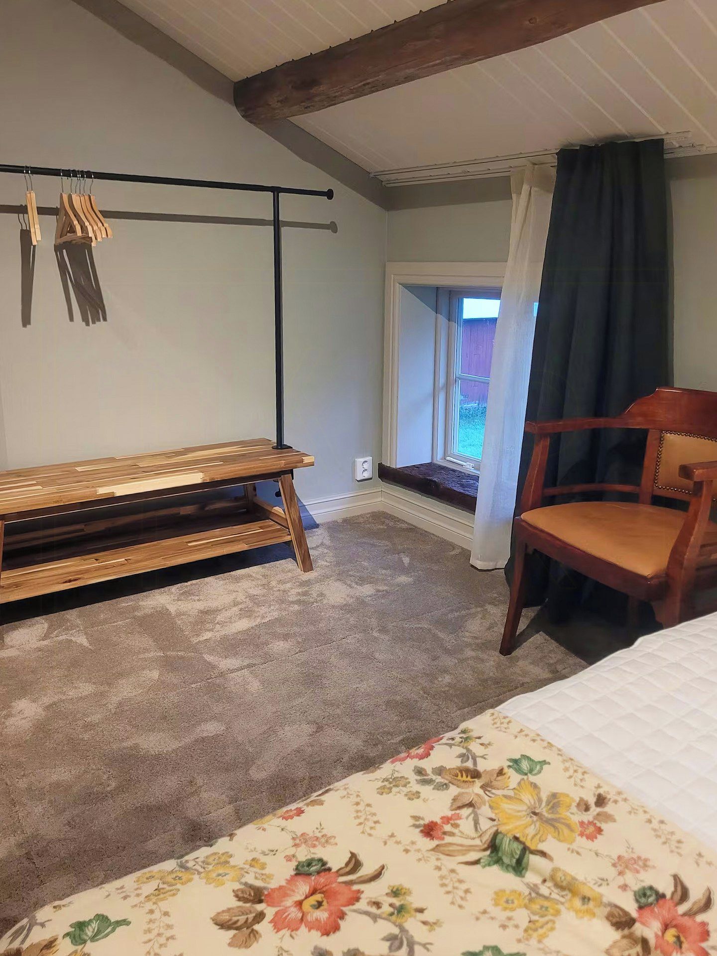 A bedroom with a wooden bench, a chair, a bed with a floral bedspread, a window with a view, and a curtain.