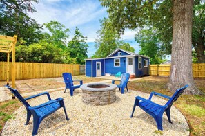 Ein Garten mit einem blauen Haus, einer Holzterrasse, einer Feuerstelle aus Stein und blauen Stühlen.