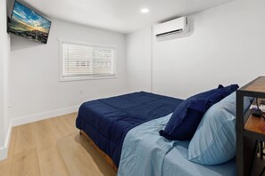 Ein Schlafzimmer mit Bett, einem Fenster mit Jalousien, einer Klimaanlage und einem an der Wand befestigten Flachbildfernseher.