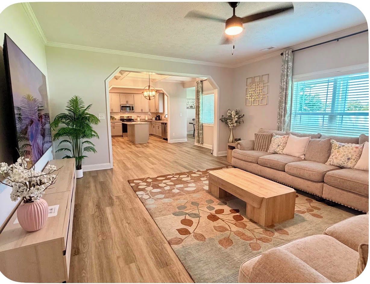 A living room with a sofa, coffee table, and a TV on a stand. There is a plant and a vase on a table.