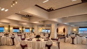 Un salón de banquetes con mesas redondas dispuestas para cenar, sillas con cintas decorativas y un amplio ventanal con vistas.