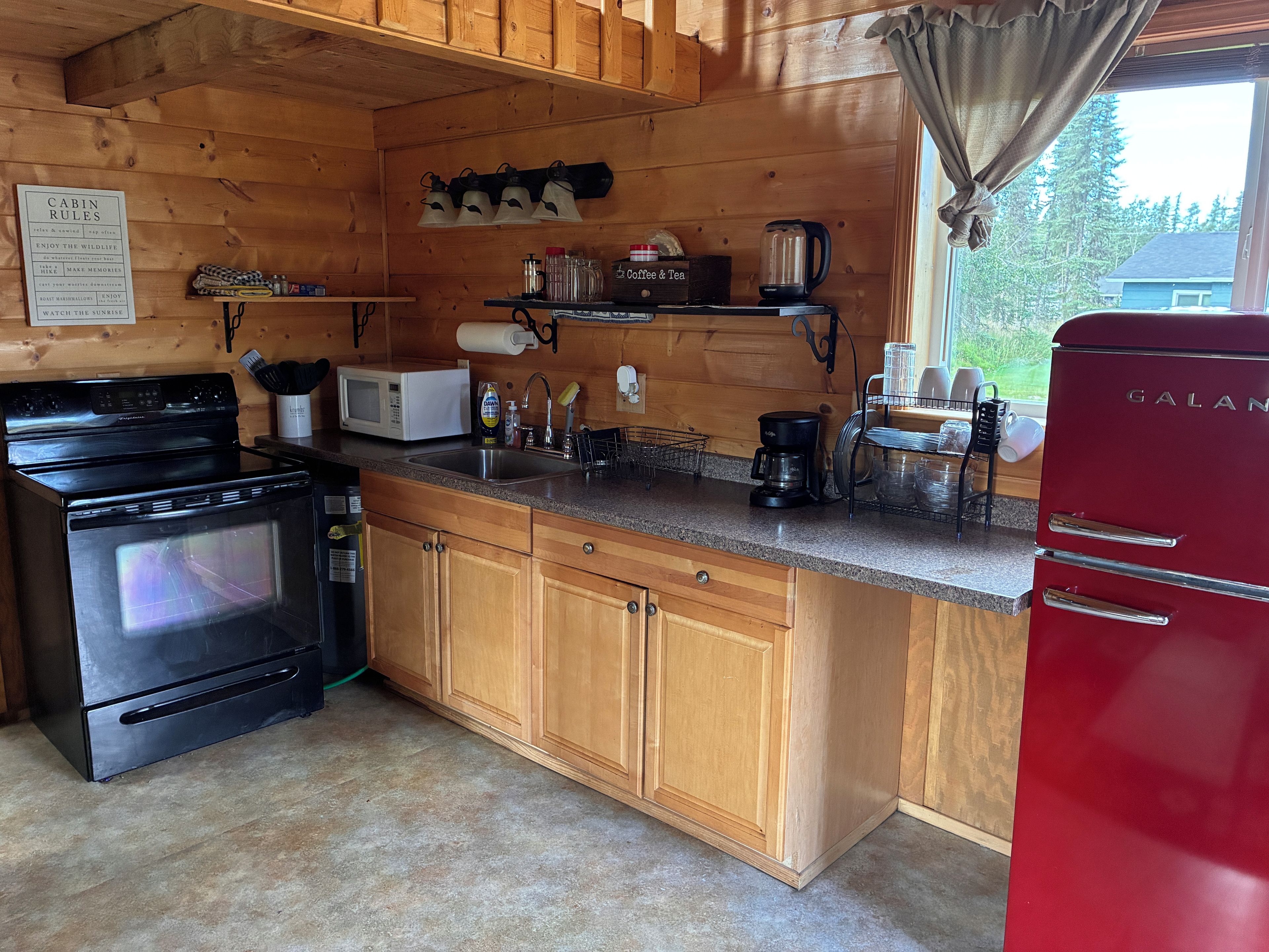 A cabin kitchen with wooden cabinets, a black stove, a microwave, a coffee maker, and a red refrigerator.