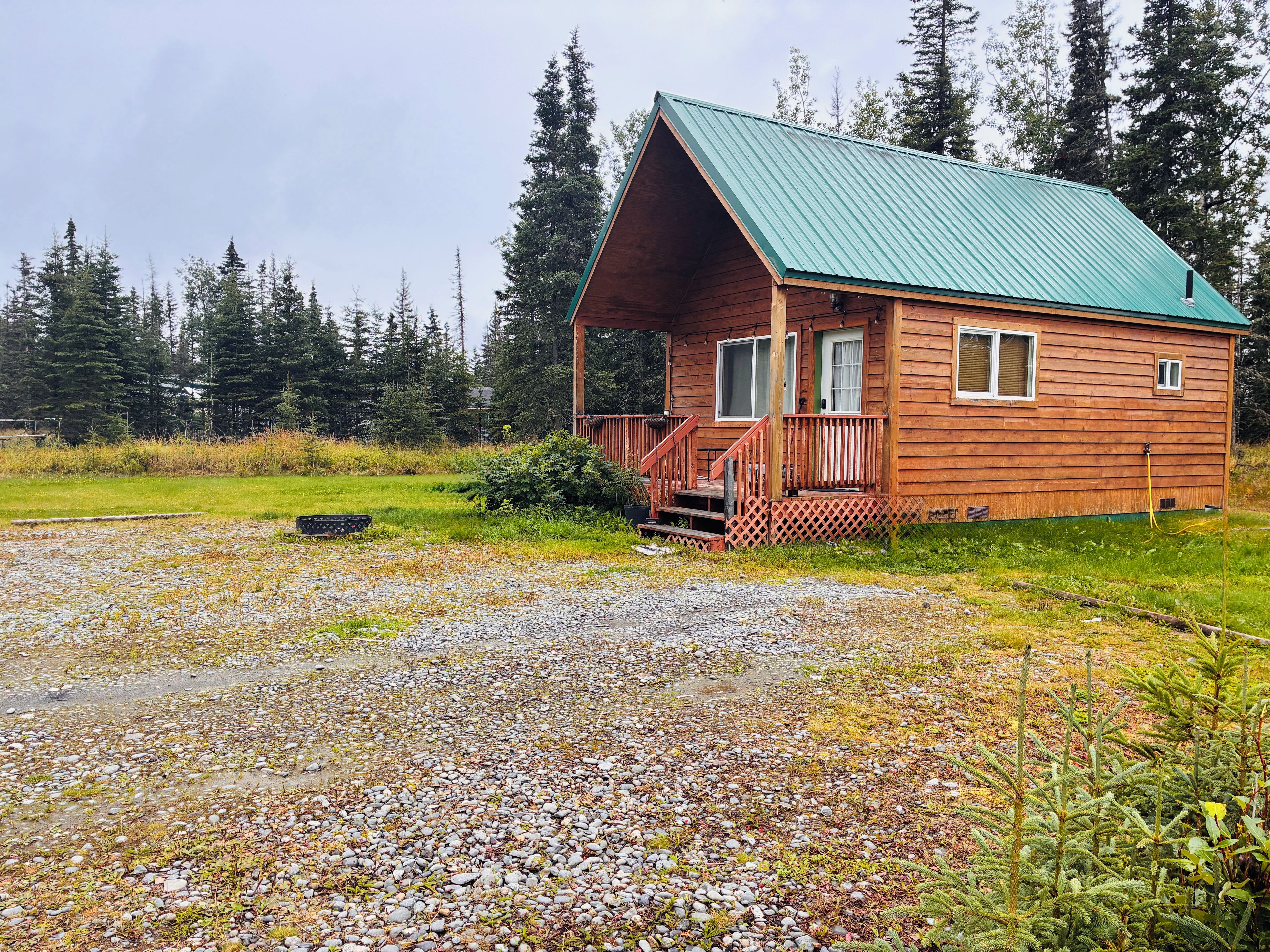 A wooden cabin with a green roof, a porch, and a gravel driveway.