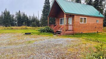 A wooden cabin with a green roof, a porch, and a gravel driveway.