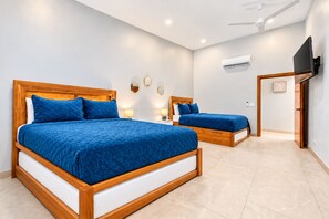 Une chambre d’hôtel avec deux lits en bois, un ventilateur de plafond, un appareil de climatisation et un téléviseur à écran plat fixé au mur.