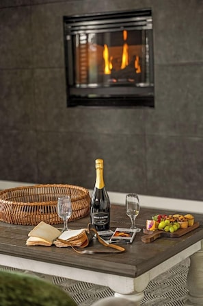 Une cheminée avec une enceinte en verre, une bouteille de champagne, deux verres, un panier, un livre et un plateau avec des fruits et des pâtisseries.