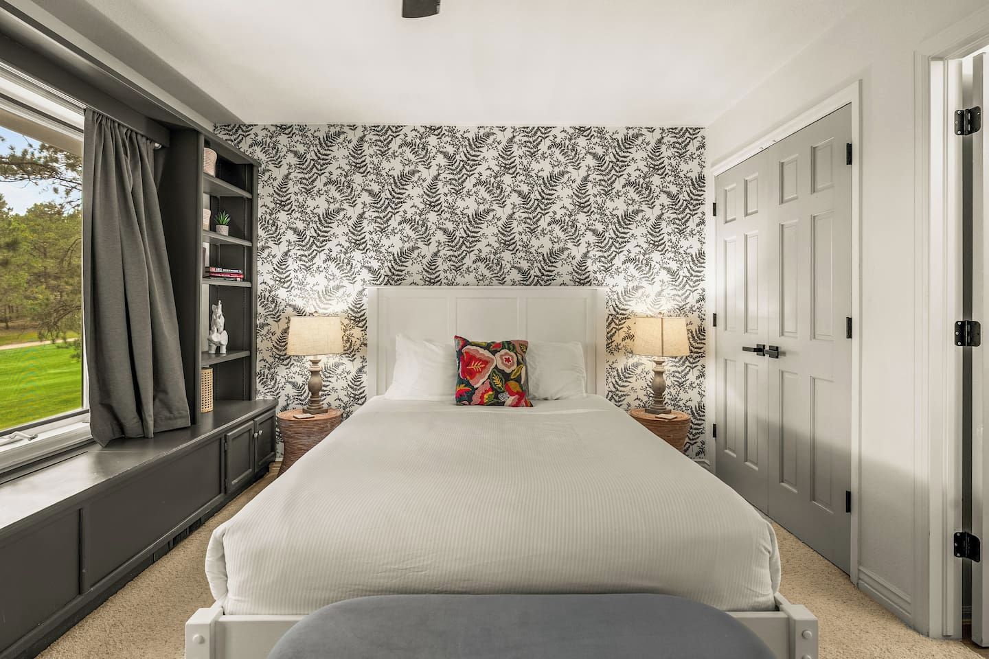 A bedroom with a large bed, two bedside tables with lamps, a bookshelf, and a window with curtains.