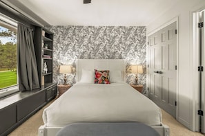 A bedroom with a large bed, two bedside tables with lamps, a bookshelf, and a window with curtains.