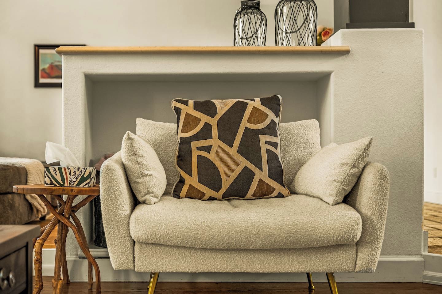 A beige armchair with a geometric patterned pillow, a small wooden side table with a tissue box, and a fireplace with decorative glass jars.