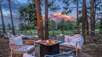 An outdoor seating area with a fire pit, lounge chairs, and a view of a forested area.