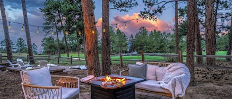 An outdoor seating area with a fire pit, lounge chairs, and a view of a forested area.
