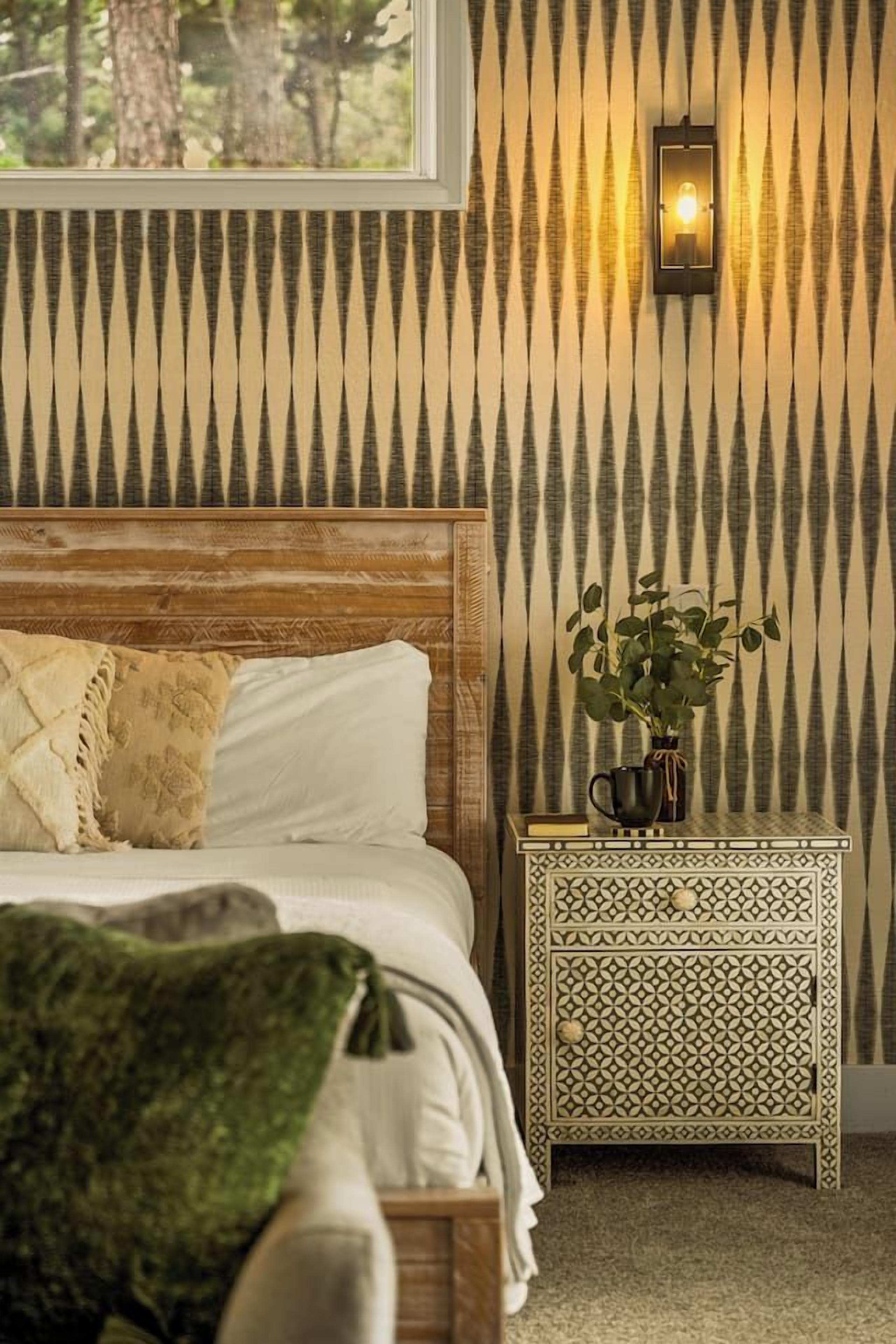 A bedroom with a wooden headboard, a bed with white linens, a patterned wall, a nightstand with a plant, and a window with a view of trees.