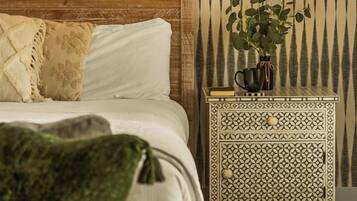 A bedroom with a wooden headboard, a bed with white linens, a patterned wall, a nightstand with a plant, and a window with a view of trees.