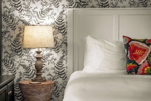 A bed with white linens and a colorful pillow, a lamp with a textured shade, and a patterned wallpaper with fern designs.